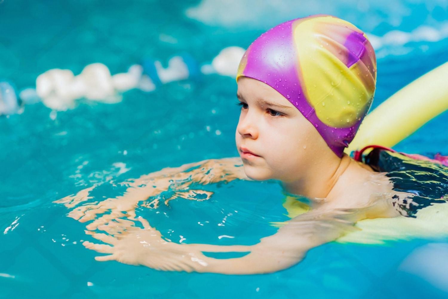 Child in a swim cap using a foam noodle to stay afloat during a school pool class